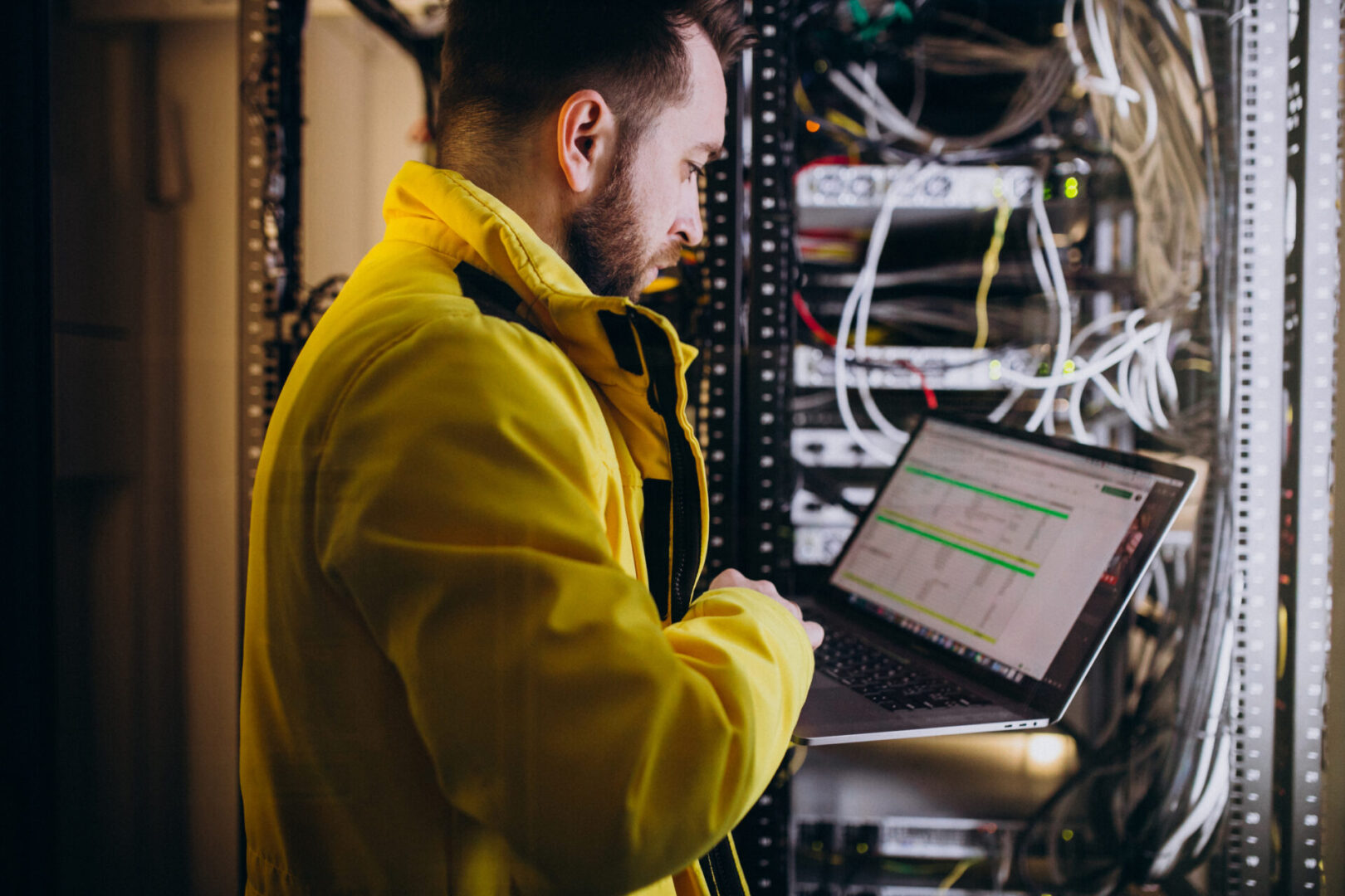 A man in yellow jacket using laptop near wires.